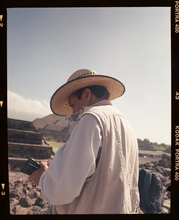 Adult male in a straw hat and white shirt is focused on a small black electronic device on the stone steps of Teotihuacan pyramid.