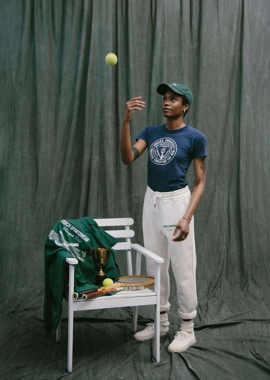 A woman in a t-shirt and sweatpants tossing a tennis ball next to a chair with tennis equipment and a trophy, with a green backdrop.