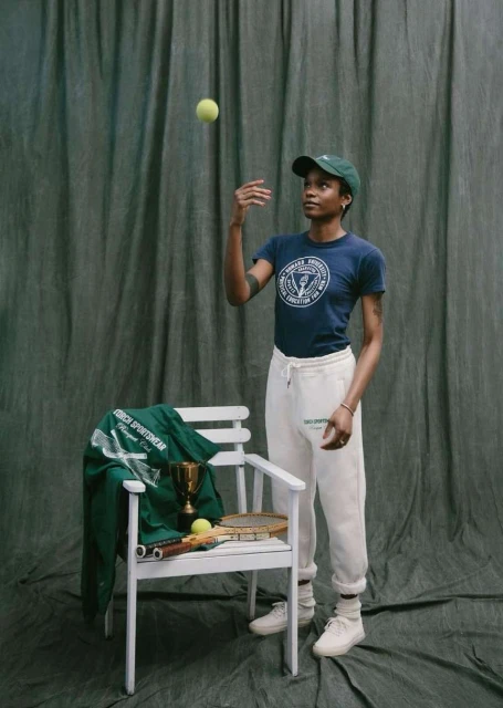 A woman in a t-shirt and sweatpants tossing a tennis ball next to a chair with tennis equipment and a trophy, with a green backdrop.