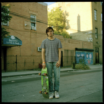Full shot of a young adult man in jeans and striped tee shirt, holding a skateboard, standing in an urban street setting with brick buildings.