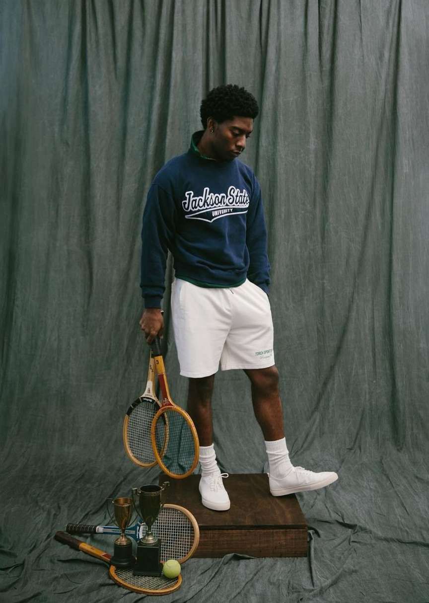 Portrait of a young Black man in athletic wear holding tennis rackets. He stands on a wood platform with tennis gear.
