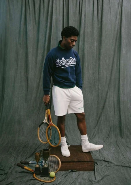 Portrait of a young Black man in athletic wear holding tennis rackets. He stands on a wood platform with tennis gear.