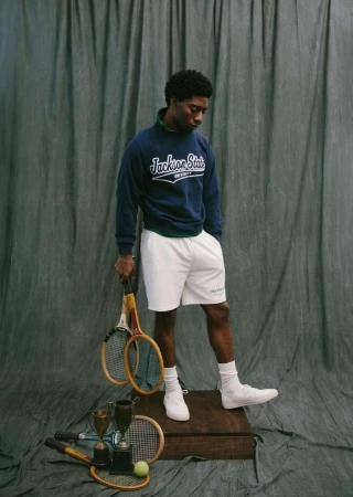 Portrait of a young Black man in athletic wear holding tennis rackets. He stands on a wood platform with tennis gear.