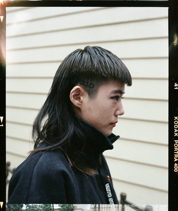Close-up profile portrait of an East Asian person with a mullet hairstyle, wearing a black jacket in front of a siding wall.