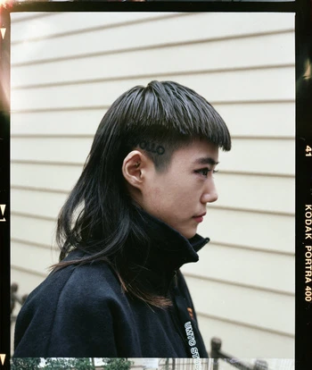 Close-up profile portrait of an East Asian person with a mullet hairstyle, wearing a black jacket in front of a siding wall.