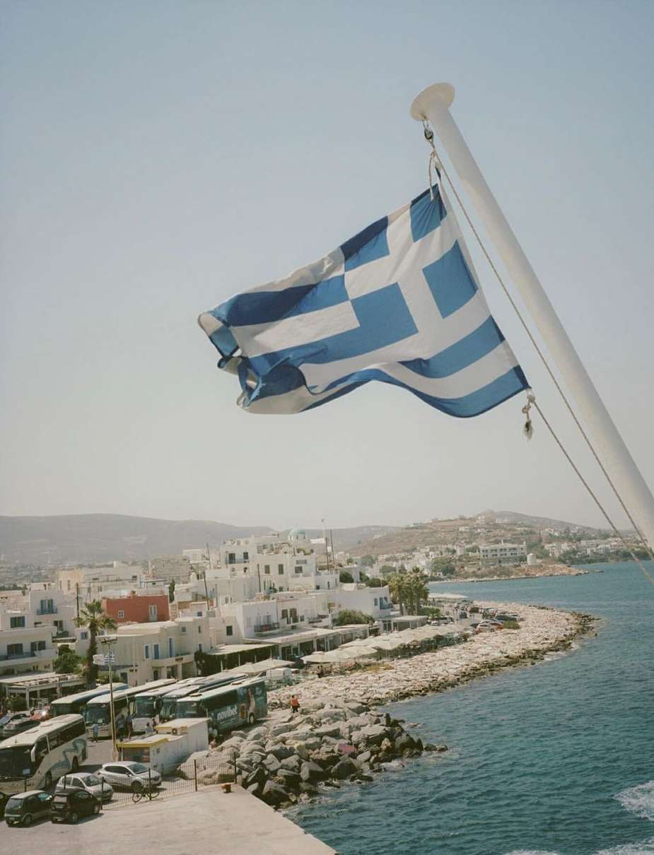 Greek flag waves above a coastal town with white buildings and parked buses along the rocky shore, on a sunny day with a clear sky.