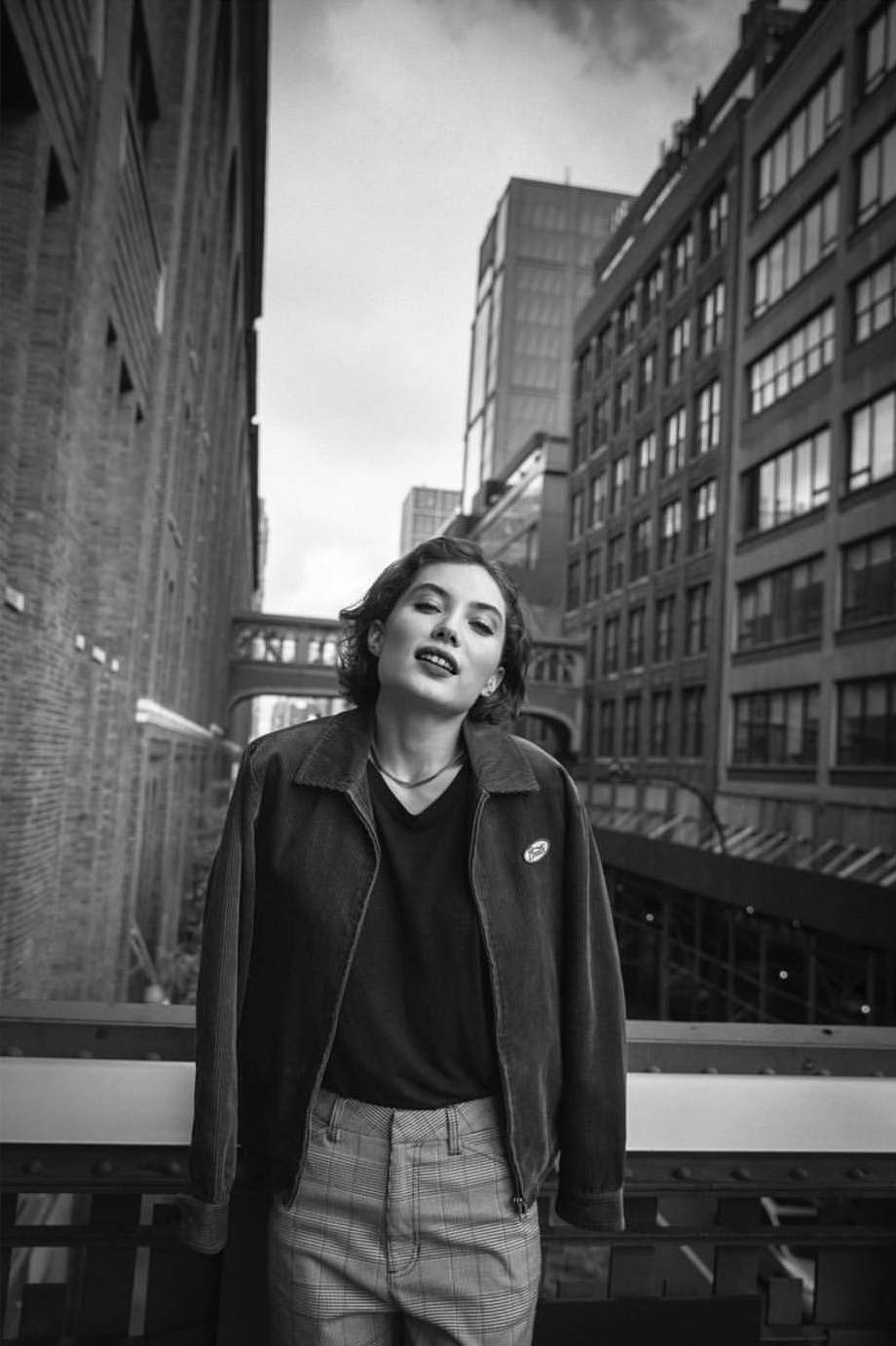 Black and white portrait of a woman in a corduroy jacket and patterned pants standing in an urban environment with tall buildings.