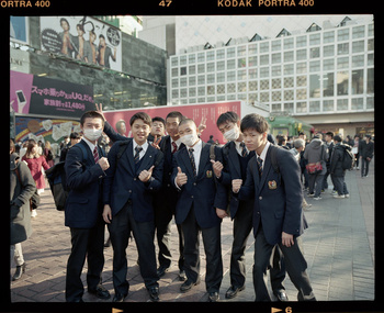 Group of Japanese high school students in school uniforms and face masks pose outdoors with city buildings and a large billboard in background.