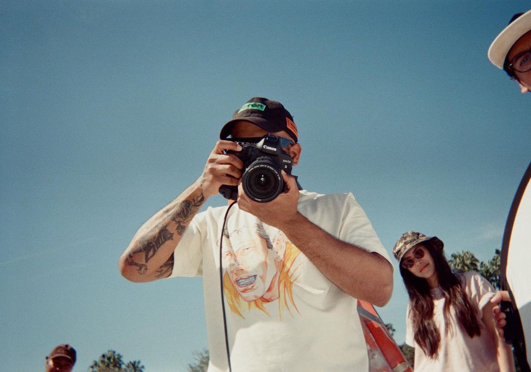 A photographer holds a camera, focusing on a subject, outdoors under a clear blue sky, with a group of people and trees visible.