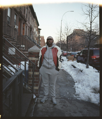 Full-body portrait of an adult person in a track suit standing on a sidewalk next to an apartment building in an urban setting.