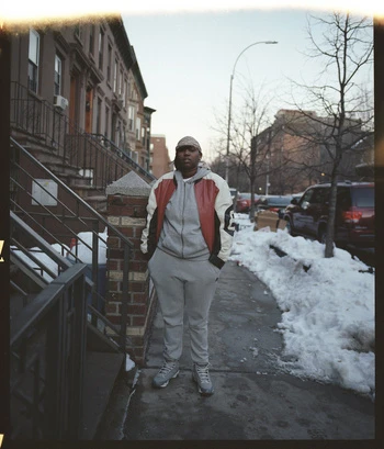 Full-body portrait of an adult person in a track suit standing on a sidewalk next to an apartment building in an urban setting.