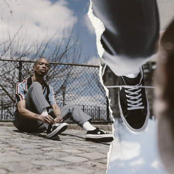 An adult man sits on a stone surface with black sneakers; ripped paper reveals a close-up shot of the black sneakers on a mirror.