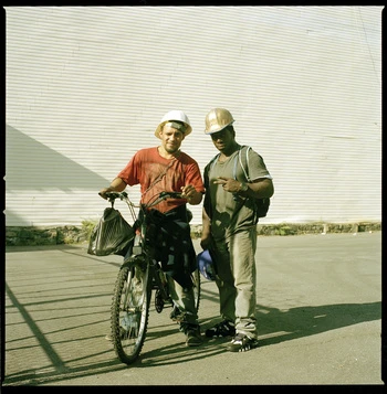 Two construction workers pose with a bicycle in front of a wall, wearing hard hats and casual clothing on a sunny day.