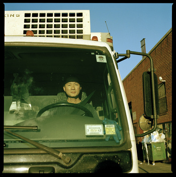 Driver in the cab of a white delivery truck on a city street on a sunny day, featuring a brick building and clear blue sky.