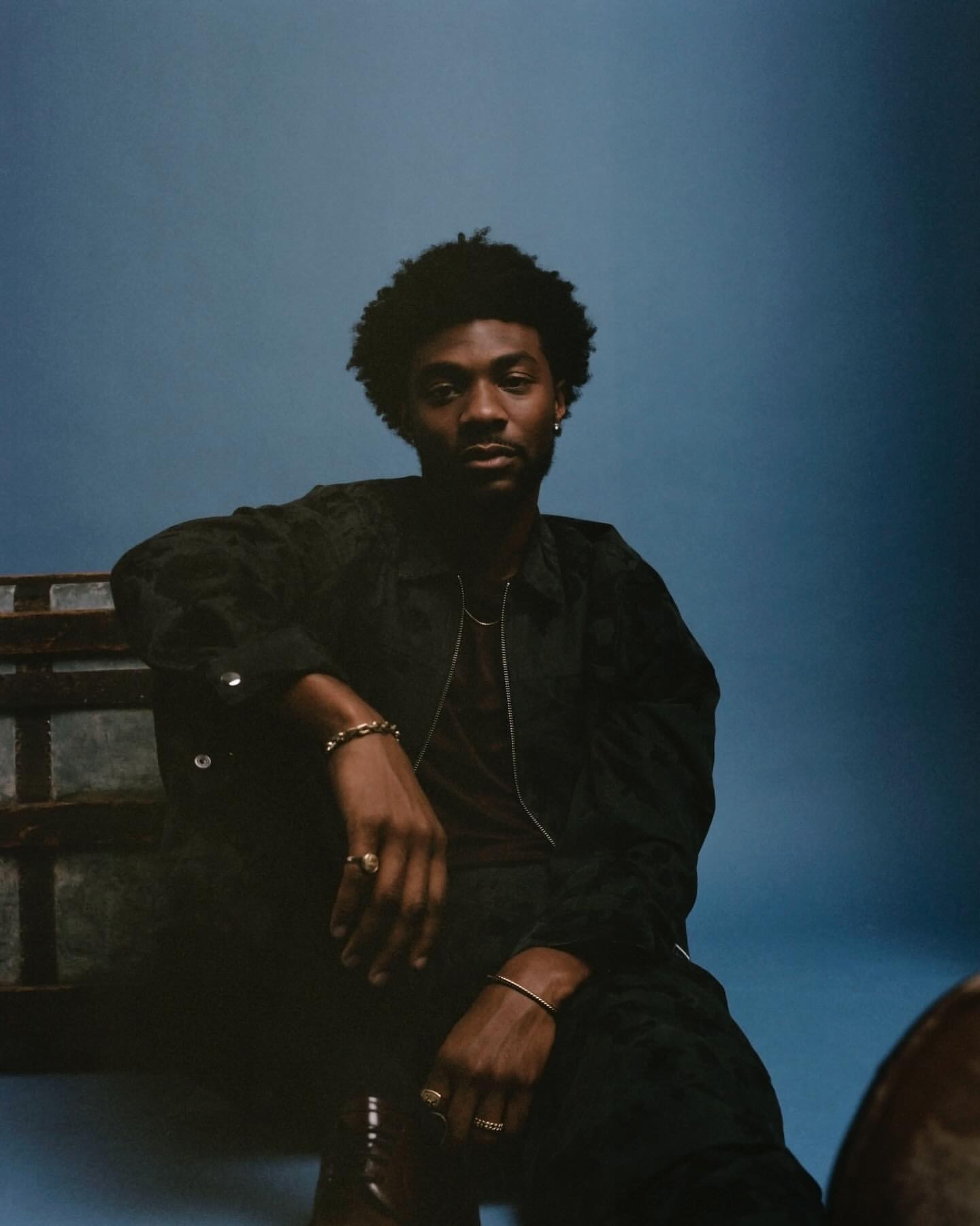 Portrait of a dark-skinned man with an afro, wearing a dark shirt and gold jewelry, sitting on an old chest against a blue backdrop.