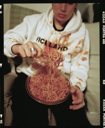 Person in a hoodie eating spaghetti from a glass bowl with a fork, some of it spilled on the white hoodie, indoor scene.