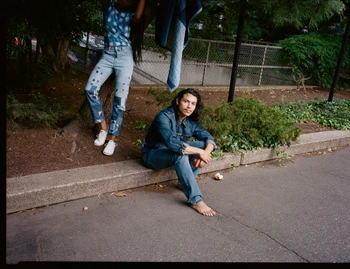 Man seated barefoot on concrete curb next to a standing woman in patchworked denim jeans, photographed outdoors by Levi Walton.