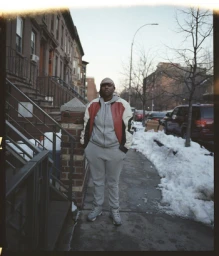 Portrait of a person wearing a patterned durag, grey hoodie, and red-and-white jacket on a snowy sidewalk.
