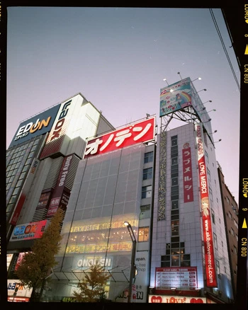 Low angle view of illuminated commercial buildings and bright neon signage against a dusk sky captured on analog film.