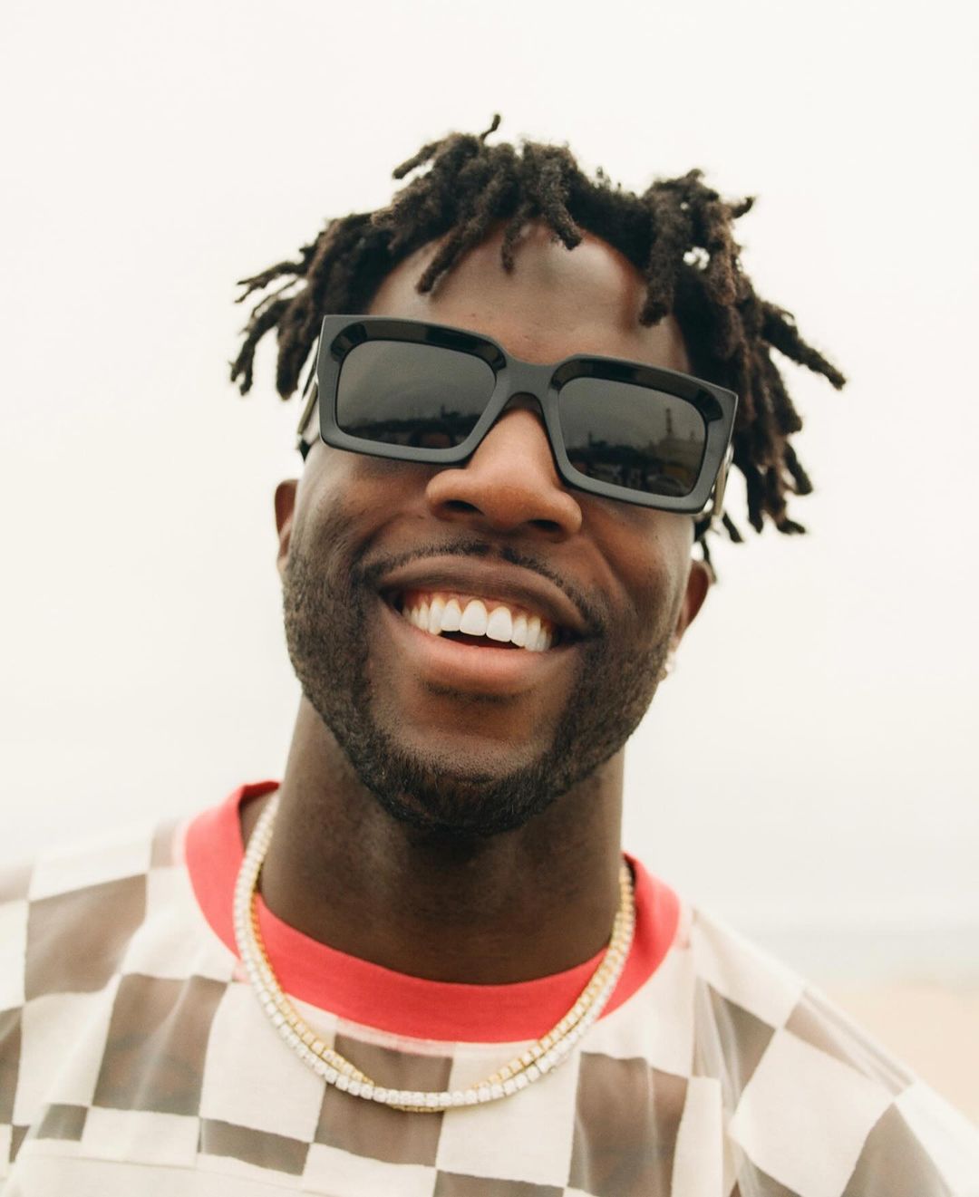 Close-up portrait of a smiling Black adult male wearing sunglasses and a gold necklace outdoors against a neutral backdrop. 