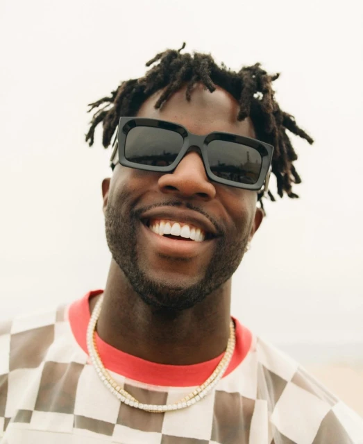 Close-up portrait of a smiling Black adult male wearing sunglasses and a gold necklace outdoors against a neutral backdrop. 