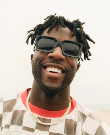 Close-up portrait of a smiling Black adult male wearing sunglasses and a gold necklace outdoors against a neutral backdrop. 