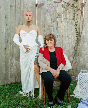 Young woman in white dress and an older woman sitting on a chair in a garden setting with flowers and a wooden fence background.