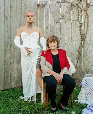Young woman in white dress and an older woman sitting on a chair in a garden setting with flowers and a wooden fence background.