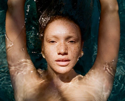Portrait of a young woman with freckles floating in turquoise water, facing the camera with raised arms and wearing gold earrings.