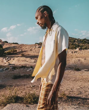 Adult male in an open shirt and plaid pants stands outdoors against a desert background with blue sky and sparse clouds.