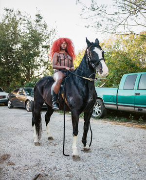 Woman with red hair riding a black horse outdoors on a gravel surface with cars and trees in the background in a medium shot.