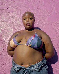 An adult woman with a shaved head poses in a colorful bikini top and jeans, standing in front of a vivid pink wall in the sunlight.