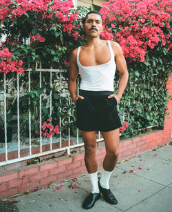Man posing in tank top and shorts in front of a brick wall and metal fence covered with pink bougainvillea flowers on the sidewalk.