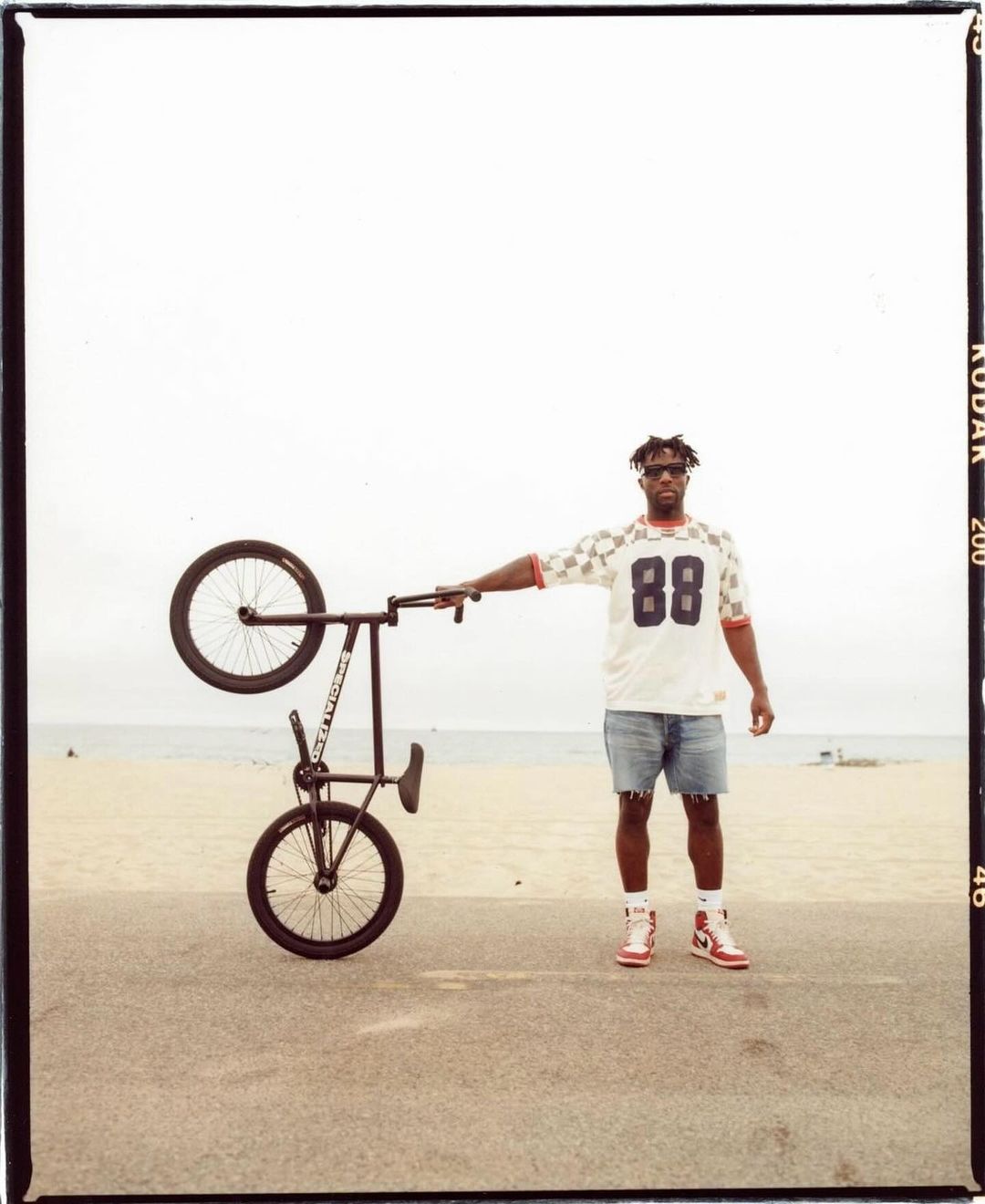 A man stands next to a black BMX bicycle held upside down on a beach, wearing a white jersey, jean shorts, red and white shoes.