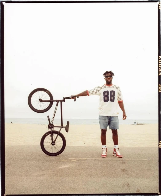 A man stands next to a black BMX bicycle held upside down on a beach, wearing a white jersey, jean shorts, red and white shoes.