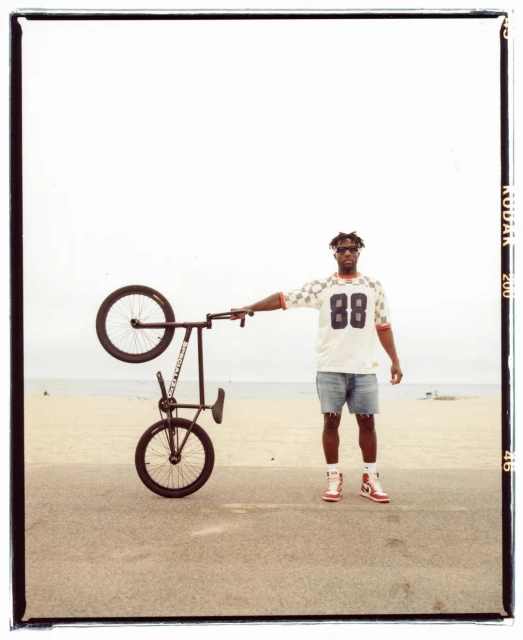 Man stands on a beach holding a BMX bicycle in the air, wearing a jersey, jean shorts, and sneakers, posing in front of the ocean.