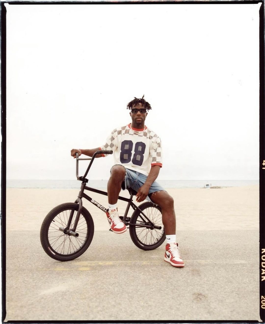 Man seated on BMX bike at beach. Adult male with sunglasses wearing retro shirt and shorts. Sandy beach and sky background.