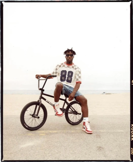Man seated on BMX bike at beach. Adult male with sunglasses wearing retro shirt and shorts. Sandy beach and sky background.