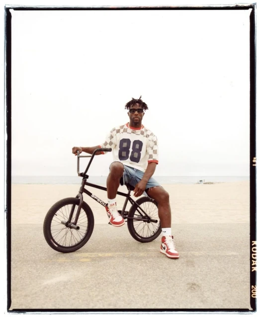 An adult male in a jersey sits on a black BMX bike outdoors on a concrete surface, wearing shorts and sneakers against a bright sky.