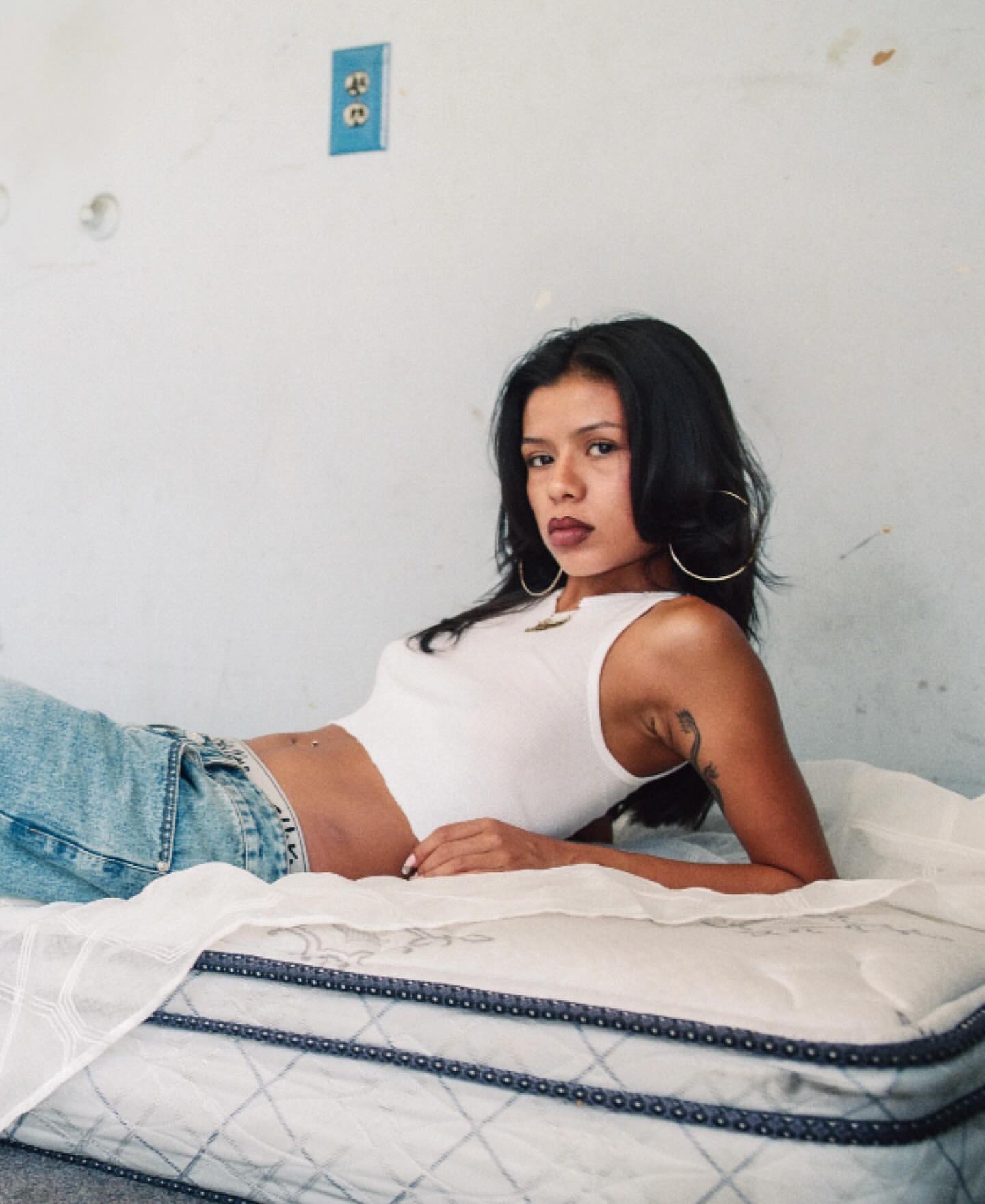 Portrait of a young woman with dark hair lying on a mattress wearing a white tank top and jeans, with a white wall in the background.