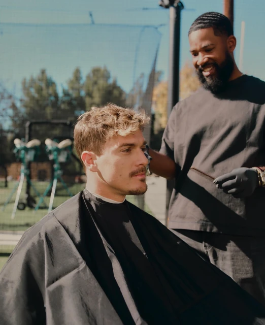 A young man getting a haircut outside from his barber, who is wearing black gloves, outdoors during the daytime on a sunny day.