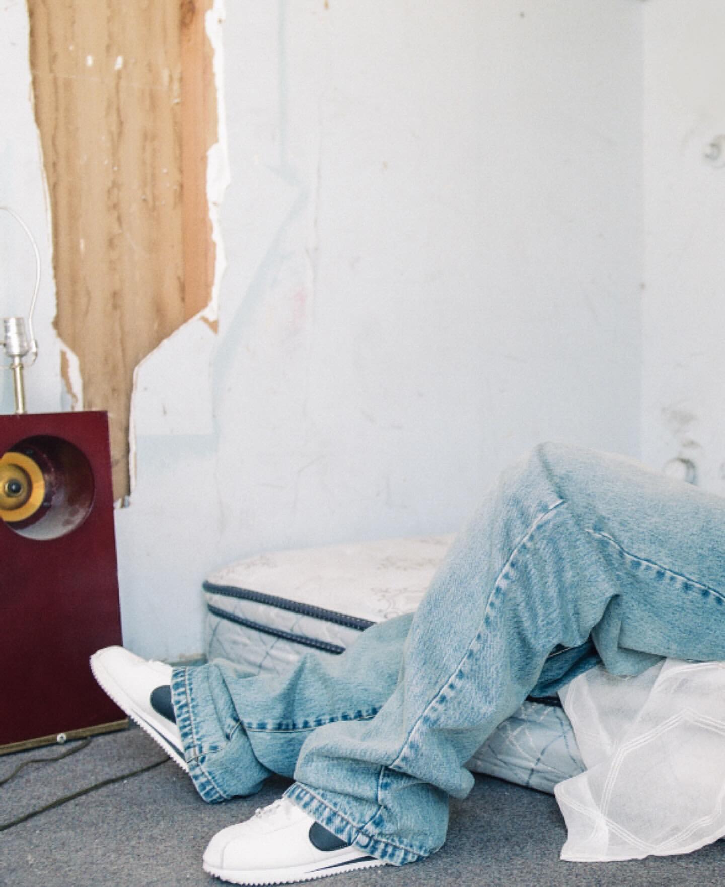 Person lying on a mattress on the floor with vintage stereo and table lamp in a distressed room. Light blue jeans and sneakers.