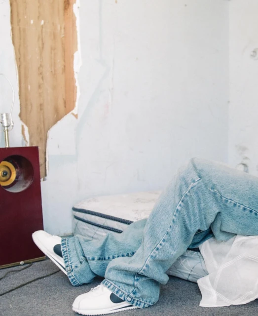 Person lying on a mattress on the floor with vintage stereo and table lamp in a distressed room. Light blue jeans and sneakers.
