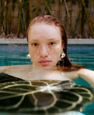 Close-up portrait of a freckled woman with wet red hair submerged in pool water, wearing metallic earrings