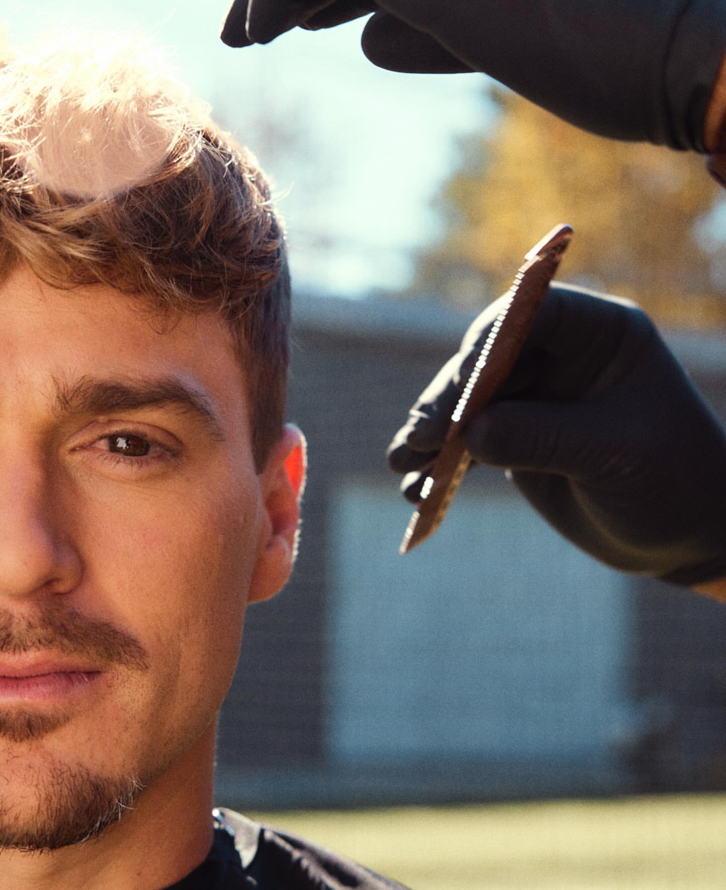 Outdoor portrait of a man getting a haircut with a comb. The barber wears black gloves. The background shows a backyard with foliage.