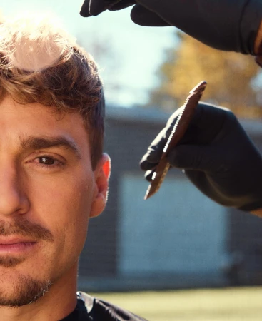 Outdoor portrait of a man getting a haircut with a comb. The barber wears black gloves. The background shows a backyard with foliage.