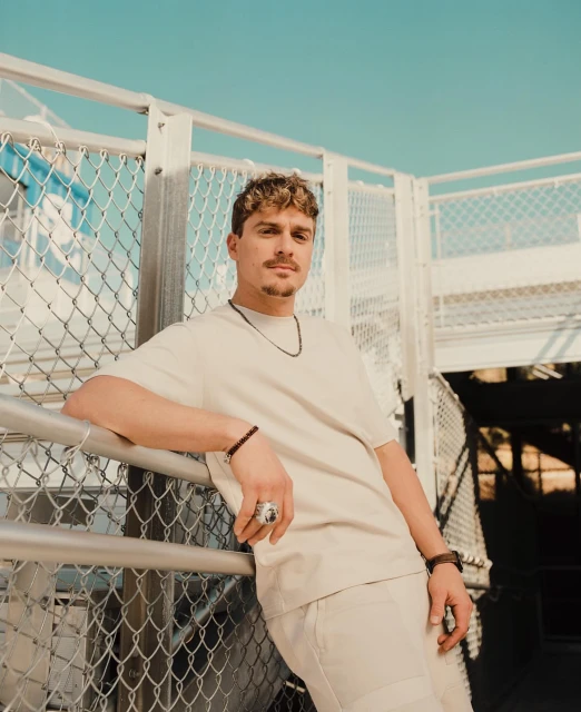 A young man with brown hair and a light complexion leans against a metal railing, wearing a tan t-shirt and matching pants under a blue sky.