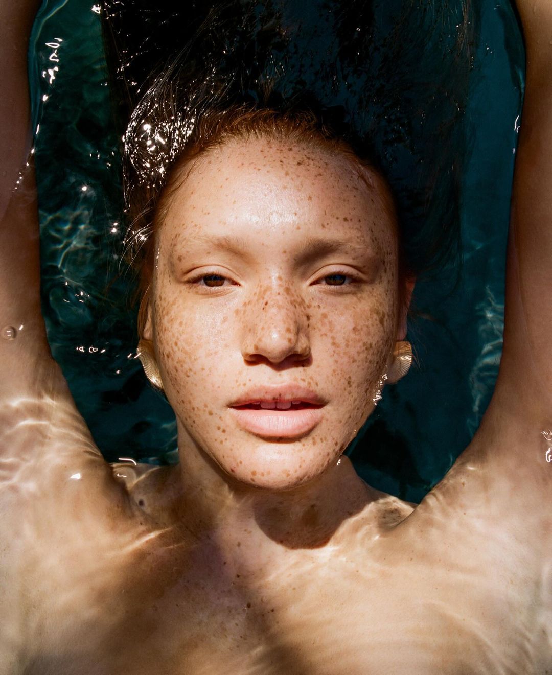Close-up of a young woman with freckles floating in blue water with dappled sunlight reflecting on the surface, looking at the camera.