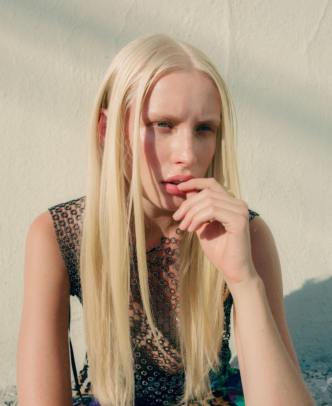Woman with long blonde hair, wearing a see-through metallic-look top, poses against a white wall in an eye-level medium shot.