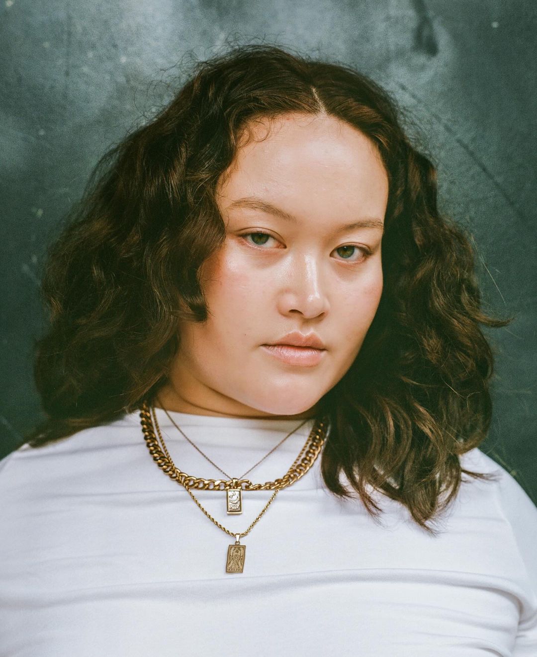 Portrait of a woman with brown hair wearing a white shirt and gold necklaces against a mottled green and grey background.